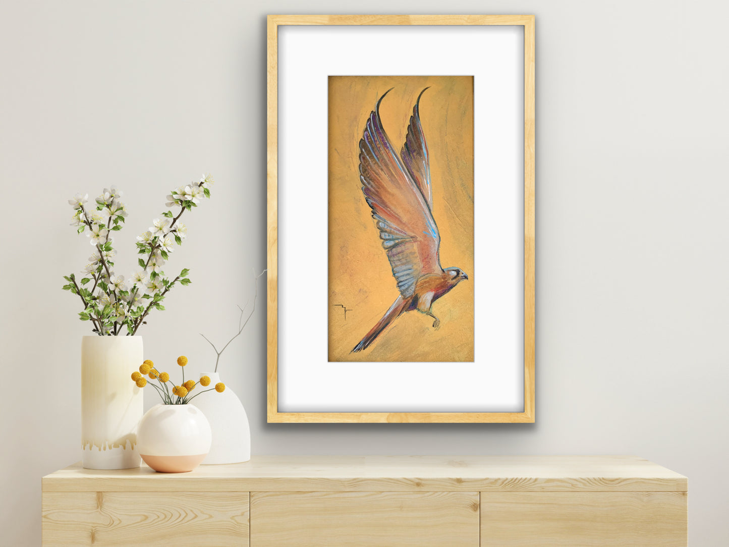'Brian' Nankeen Kestrel fine art print by Marina Fox, framed in natural timber, displayed above a light oak sideboard with white ceramic vases and white blossom branches against a soft grey wall