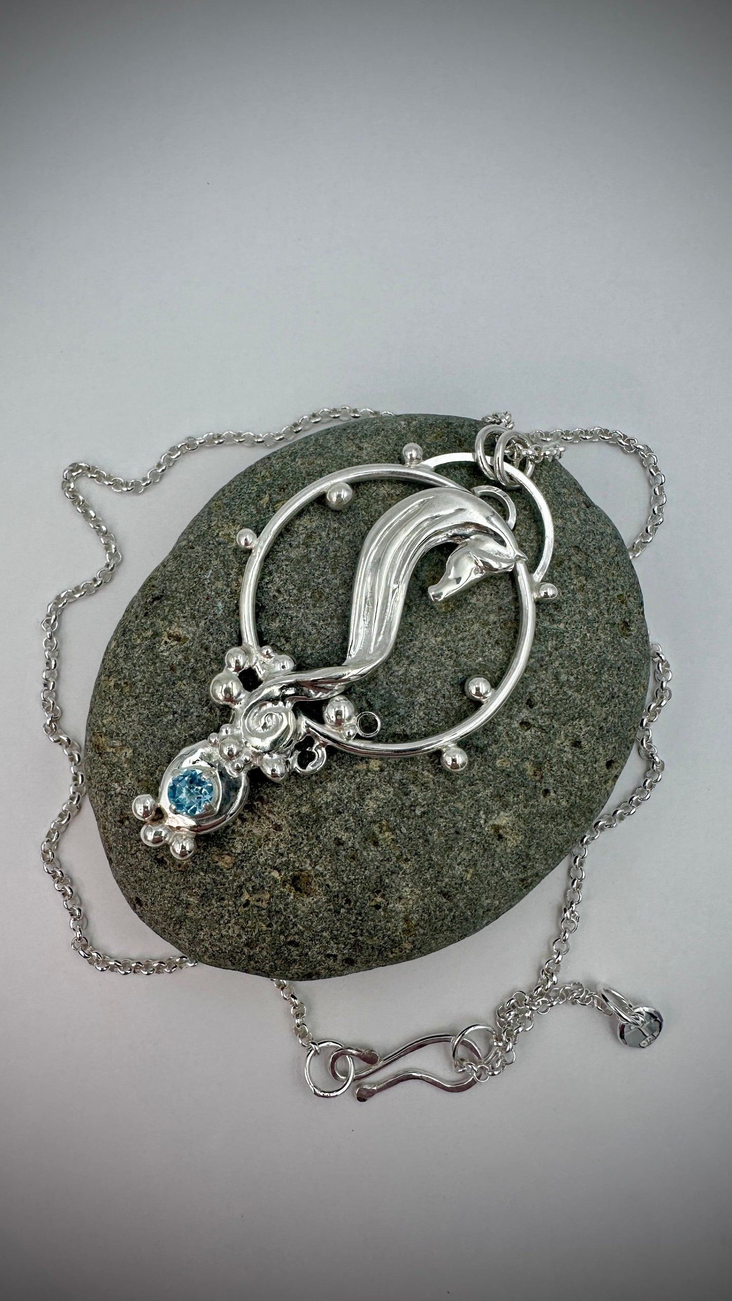 Handcrafted sterling silver seahorse pendant lying on a dark stone, with blue topaz gemstone, decorative silver balls, and ornate filigree scrollwork