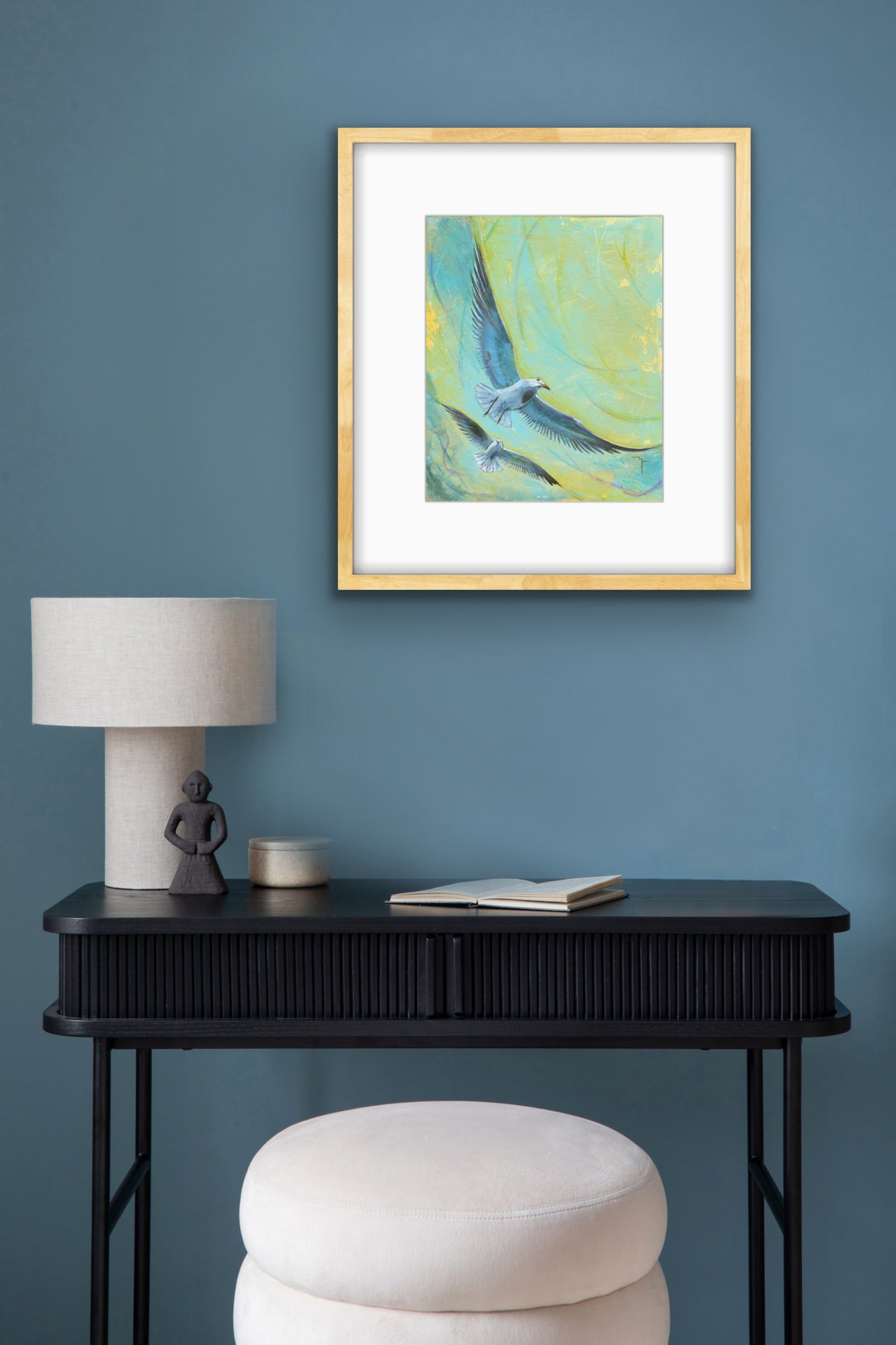 'Hide the Chips' Seagull fine art print by Marina Fox, framed in gold timber, displayed above a black ribbed console table with a lamp and figurine against a deep teal wall
