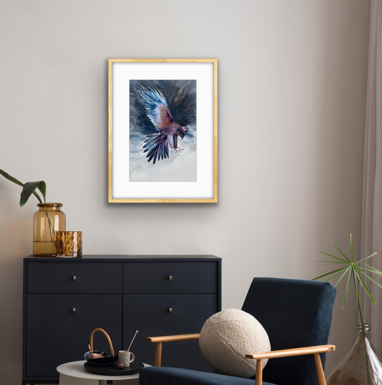 'Aerial Fury' Brown Goshawk fine art print by Marina Fox, framed in natural timber, displayed above a dark navy chest of drawers with amber glass vases beside a navy armchair in a warm neutral living room