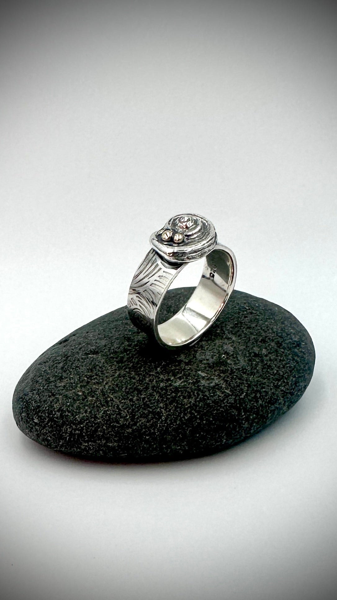 Angled view of sterling silver Beach Ring on dark river stone, showing spiral shell feature, silver granule detail, wave-textured band and polished interior — Marina Fox Fine Art