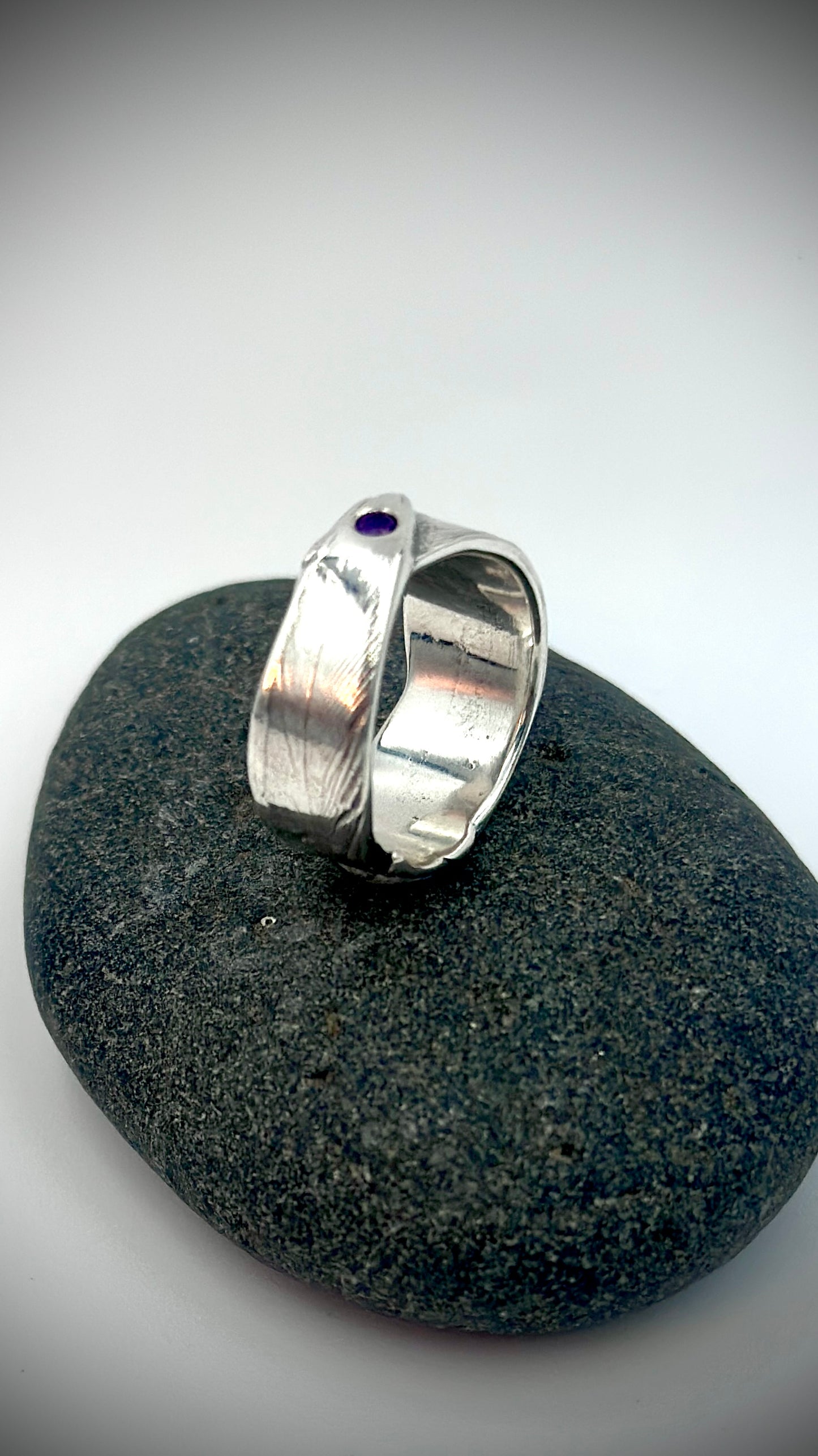 Angled view of sterling silver bird's feather ring resting on dark river stone, flush-set amethyst at crown with detailed feather texture across the overlapping band — Marina Fox Fine Art