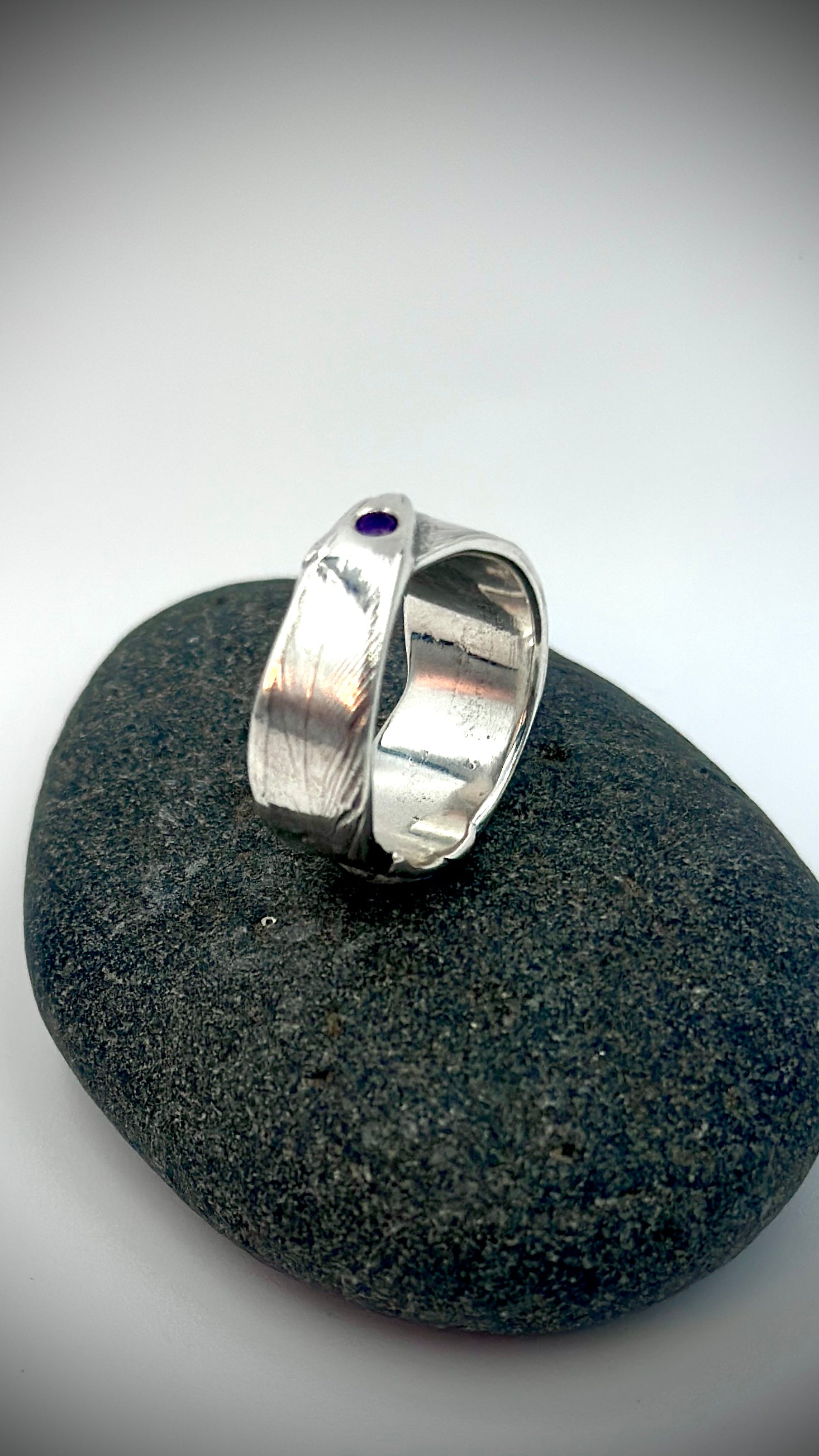 Angled view of sterling silver bird's feather ring resting on dark river stone, flush-set amethyst at crown with detailed feather texture across the overlapping band — Marina Fox Fine Art