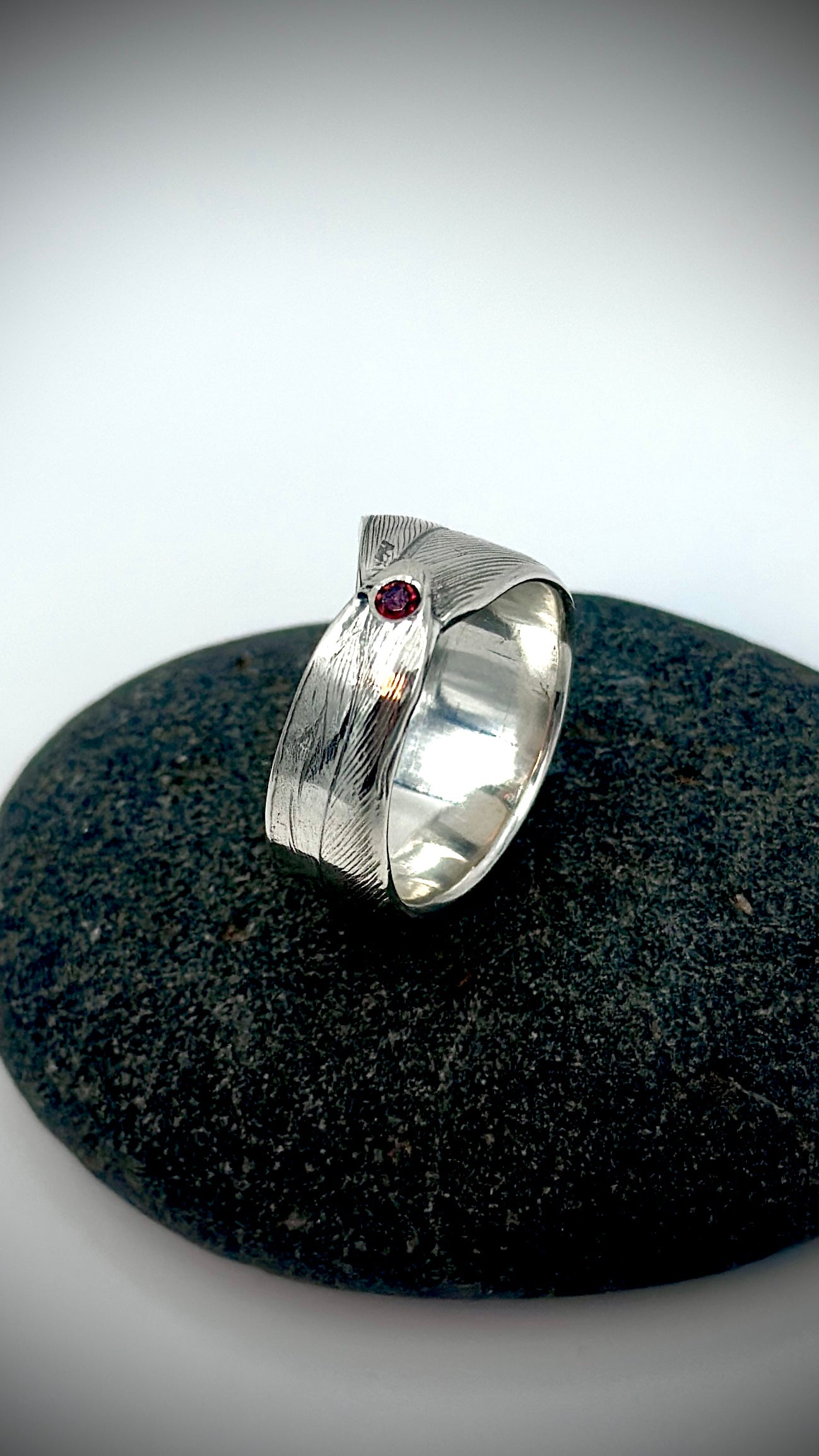 Angled view of sterling silver feather ring on dark river stone, highlighting the red garnet setting and overlapping feather band with polished interior — Marina Fox Fine Art
