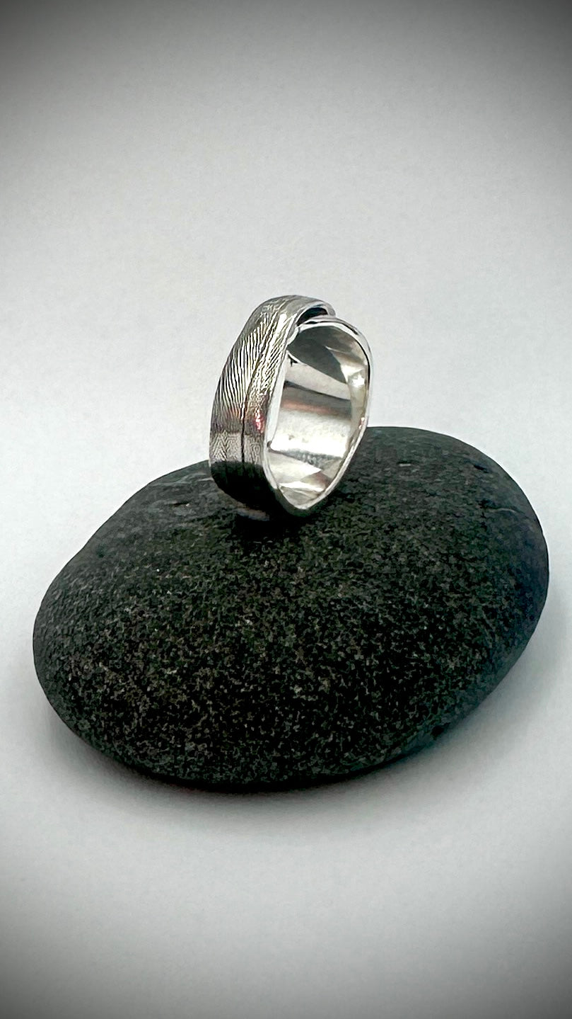 Angled view of sterling silver feather ring standing on dark river stone, showing fine feather barb texture on outer band — Marina Fox Fine Art