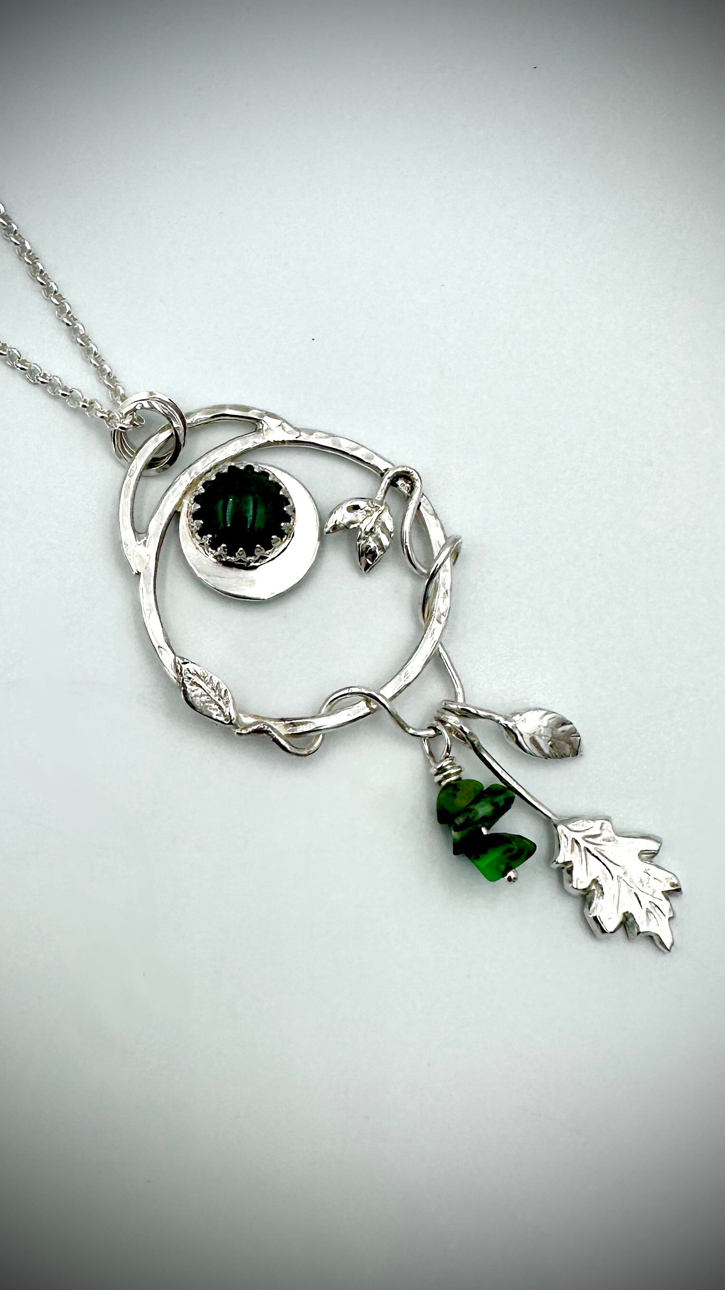 Close-up of sterling silver Dryad pendant showing green aventurine cabochon in crown setting, crescent moon, entwined branch hoop, and dangling green gemstone drop and oak leaf charms