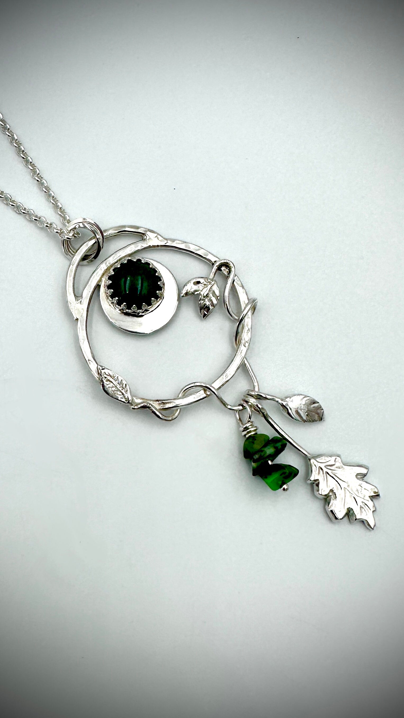 Close-up of sterling silver Dryad pendant showing green aventurine cabochon in crown setting, crescent moon, entwined branch hoop, and dangling green gemstone drop and oak leaf charms