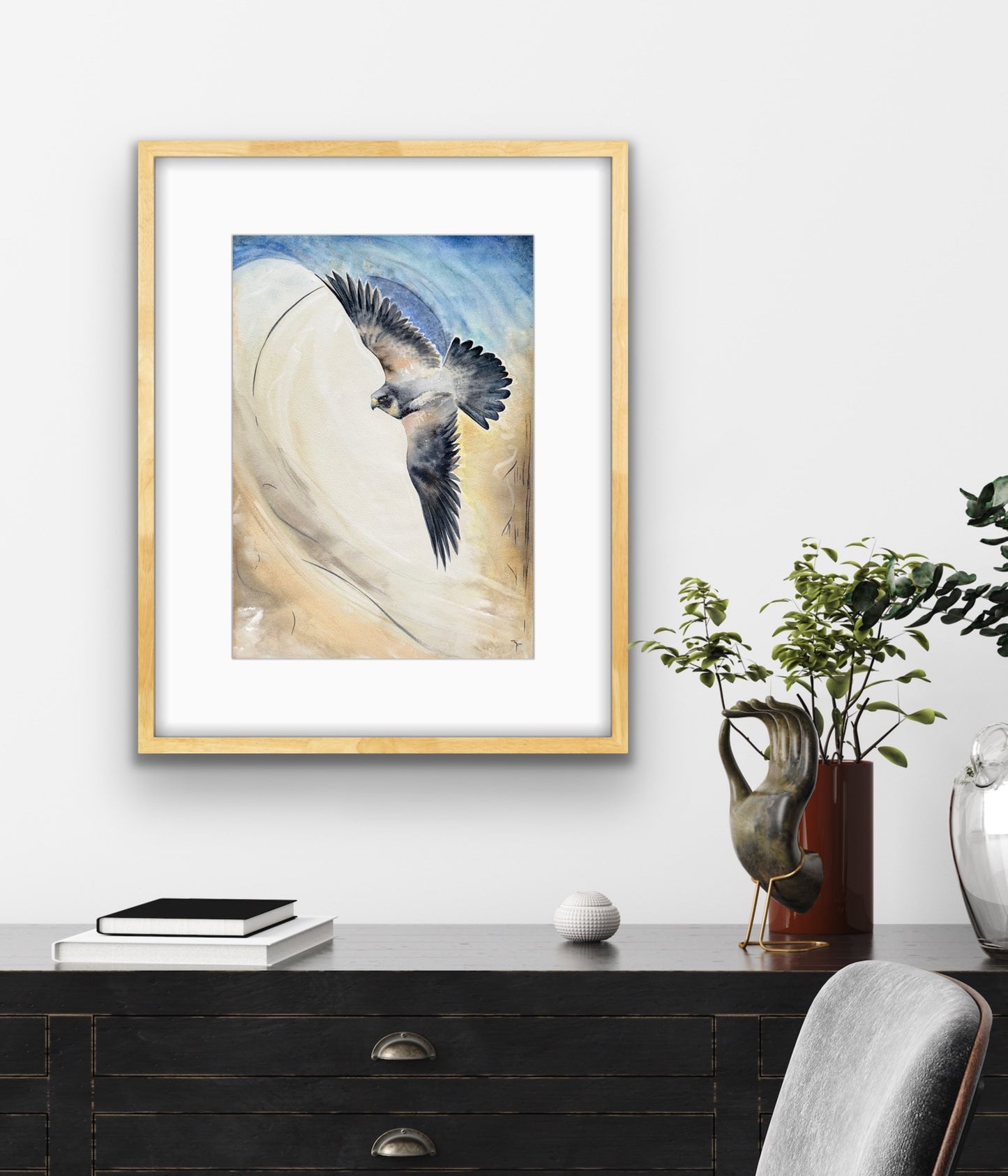 'Crosswind' Peregrine Falcon fine art print by Marina Fox, framed in natural timber, displayed above a dark timber console with greenery in a contemporary home office