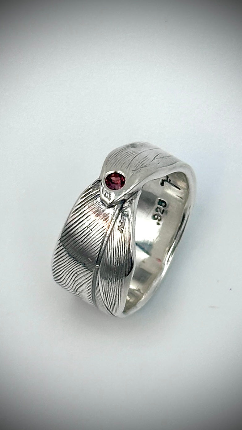 Front view of sterling silver bird's feather ring with flush-set red garnet, showing fine lost wax cast feather barb detail across the wide band — Marina Fox Fine Art