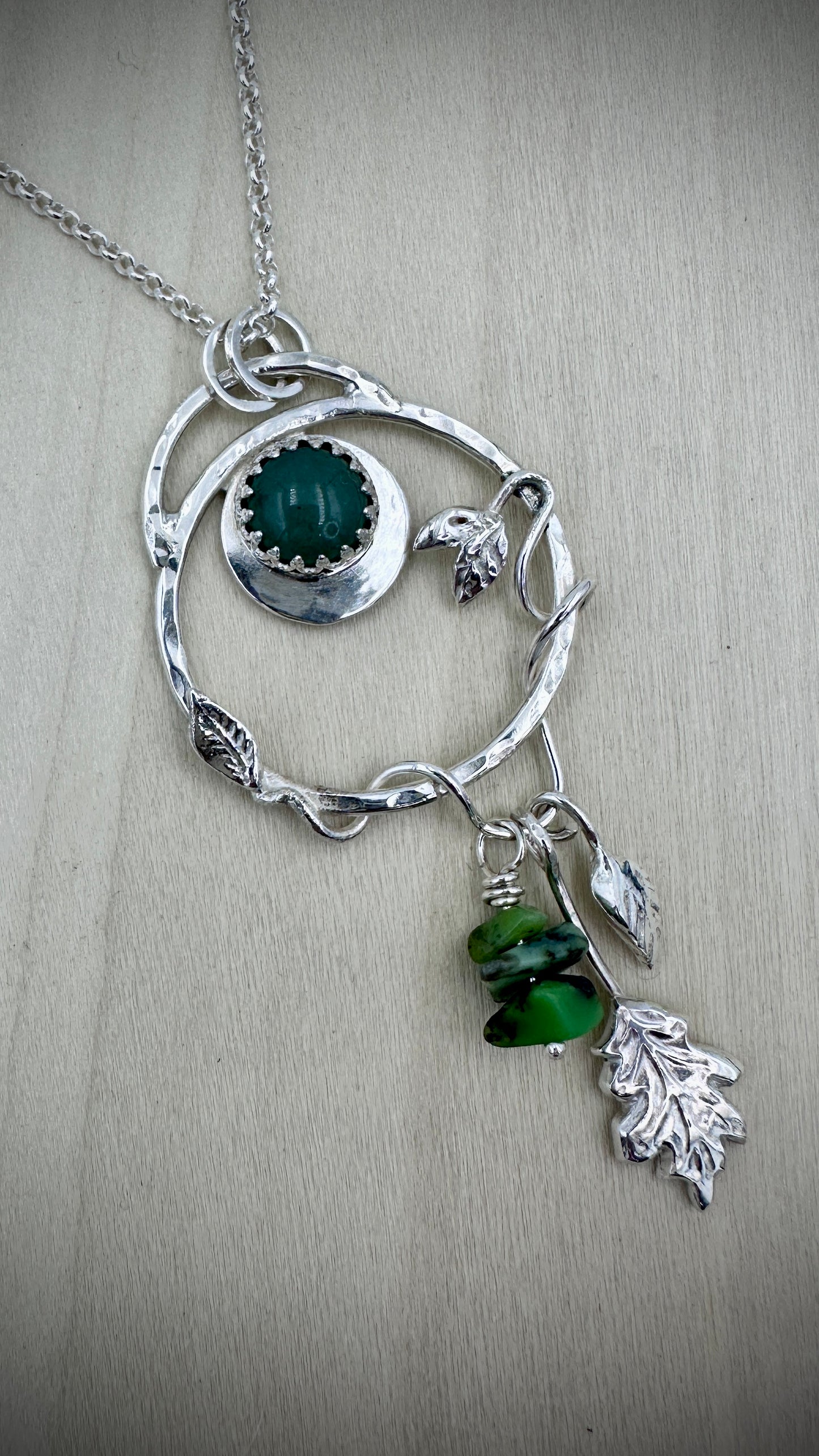 Sterling silver nature-inspired pendant necklace on a silver chain, front view on a light wood surface, showing an organic twig-form circular frame with a crown-bezel green adventurine cabochon, silver leaf and bird accents, and a dangling drop of stacked green gemstone chips with a sculpted silver oak leaf charm