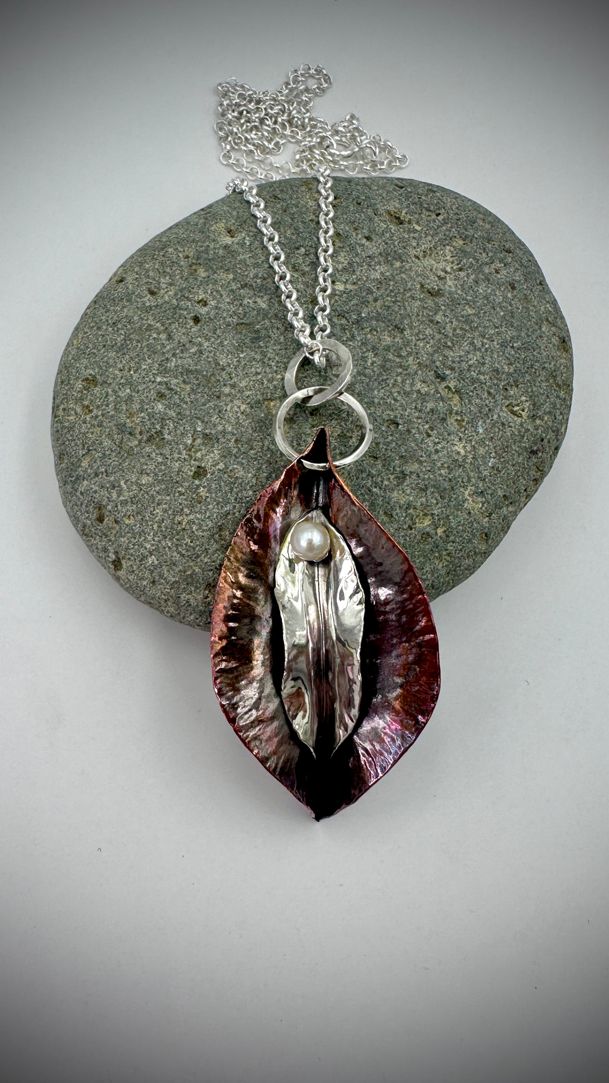 Hammered copper and sterling silver yoni pendant with freshwater pearl resting on grey river stone, white background