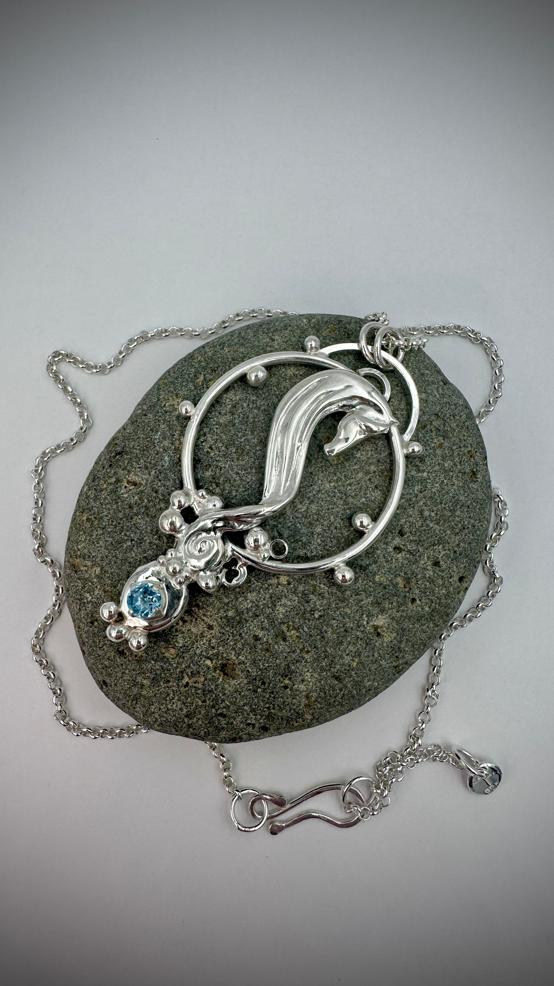 Handcrafted sterling silver seahorse pendant lying on a dark stone, with blue topaz gemstone, decorative silver balls, and ornate filigree scrollwork