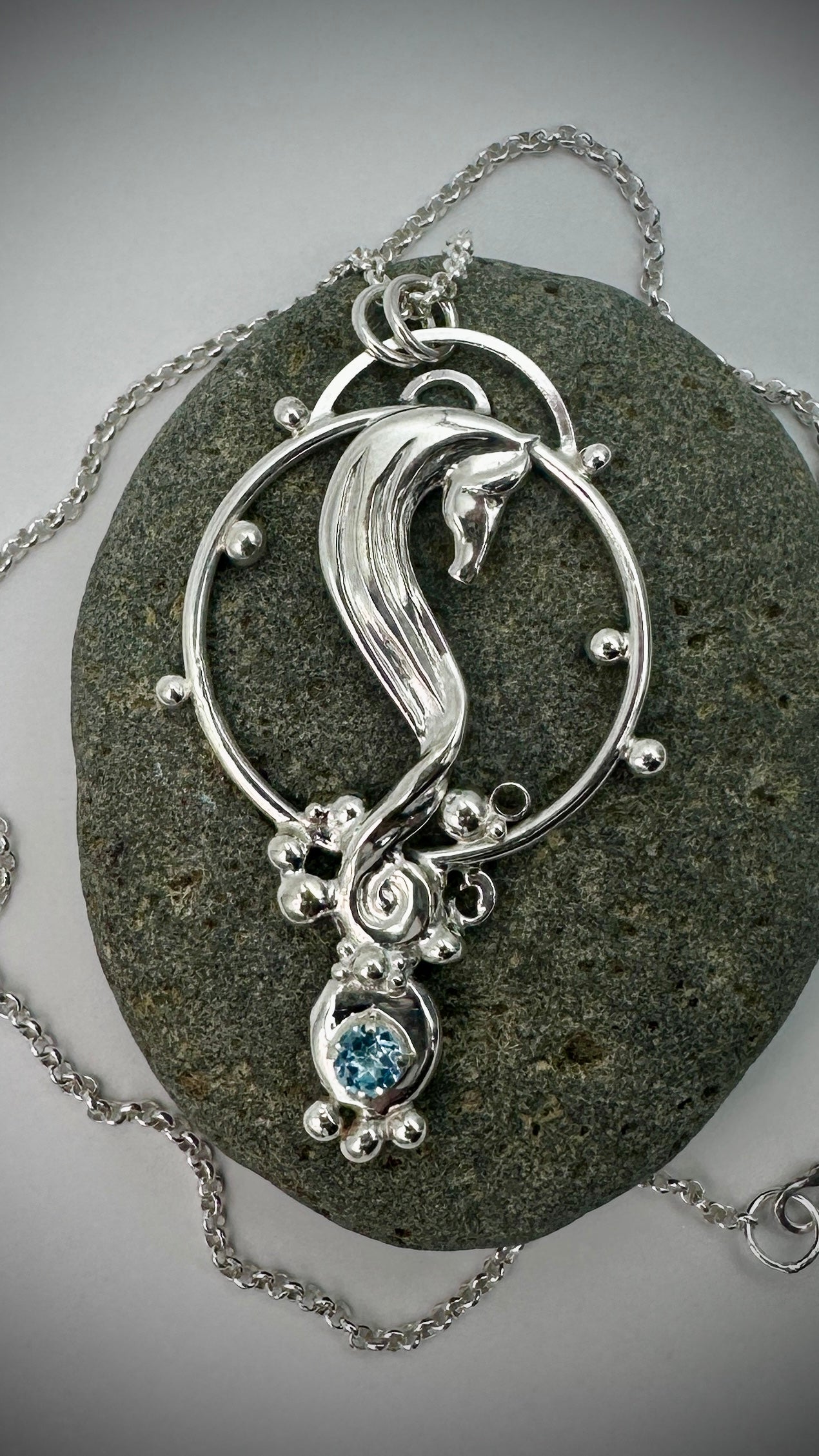 Overhead view of sterling silver seahorse pendant on a textured stone, showing full circular frame, flowing seahorse form, and blue topaz gemstone setting