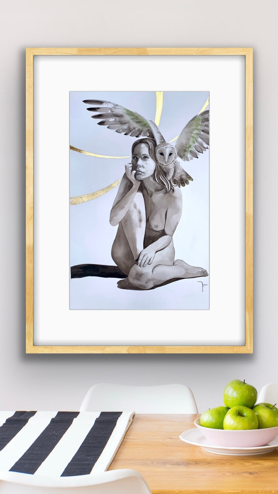 'Owl Thoughts' female figure and barn owl fine art print by Marina Fox, framed in natural timber, displayed above a timber dining table with green apples and a striped placemat against a soft grey wall