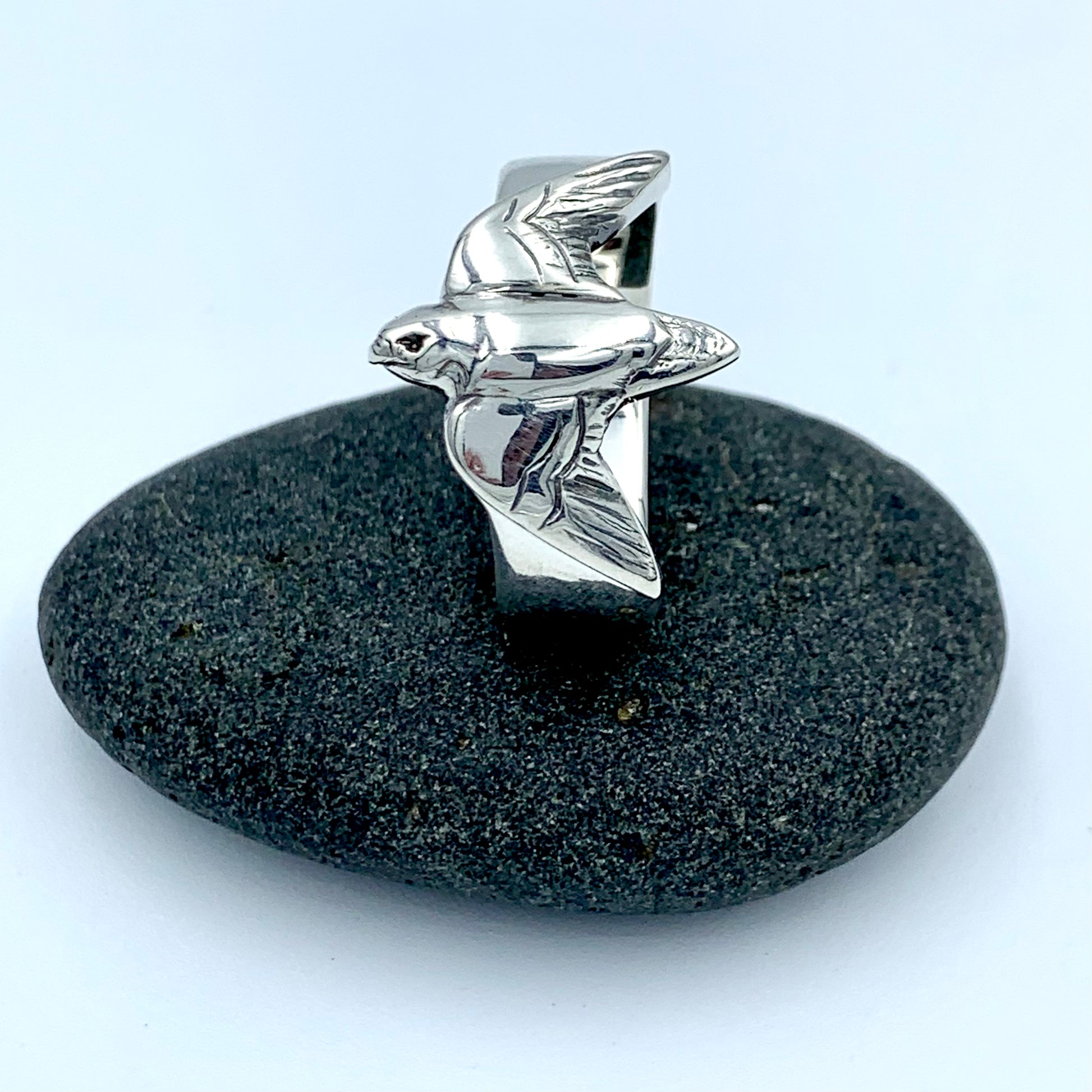 Sterling silver falconer's ring with sculpted bird in flight, angled front view on a dark river stone, showing detailed wing and body form with polished finish