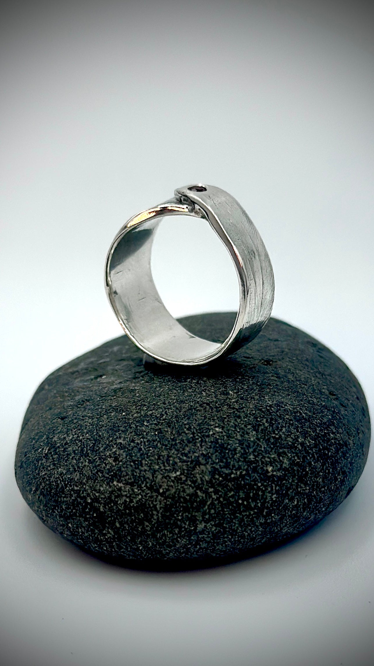 Side profile of sterling silver feather ring standing upright on dark river stone, open band form with garnet gemstone at the crown — Marina Fox Fine Art