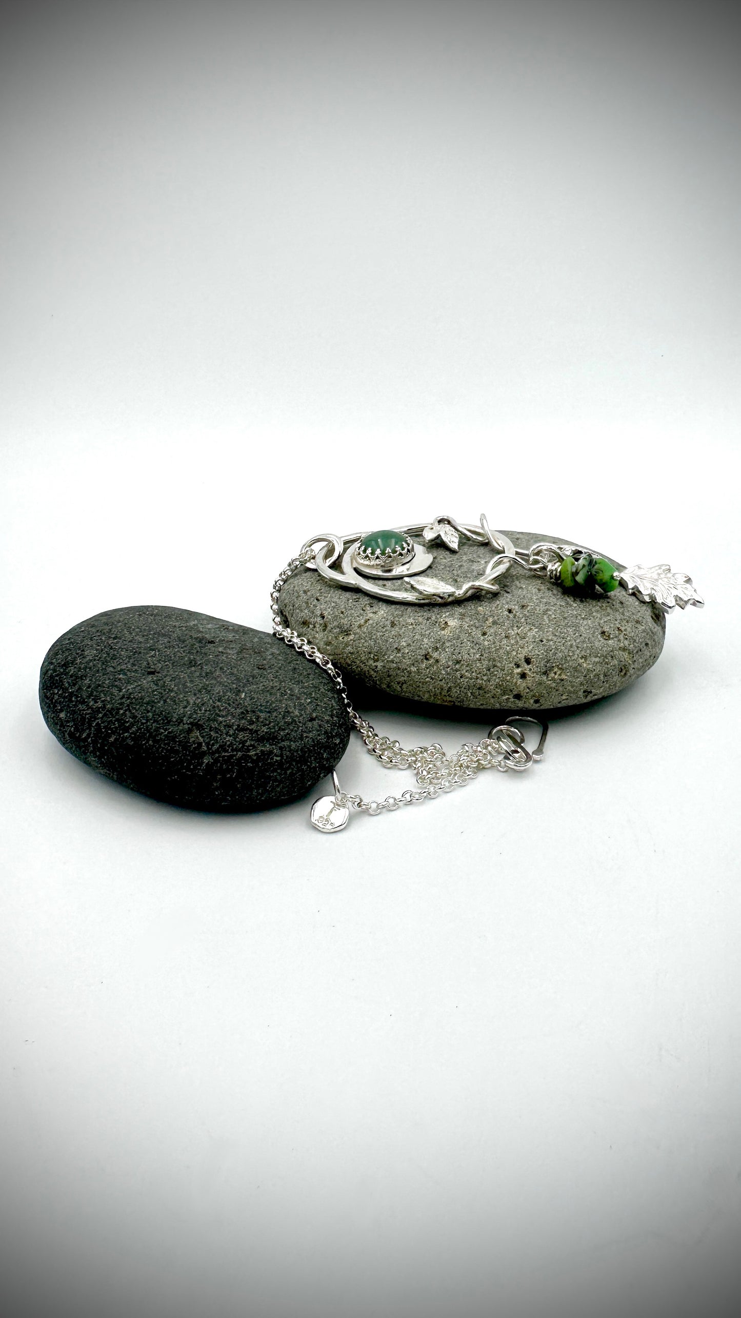 Sterling silver Dryad pendant laid flat on dark river stone with chain extended, showing botanical leaf and oak charm drops