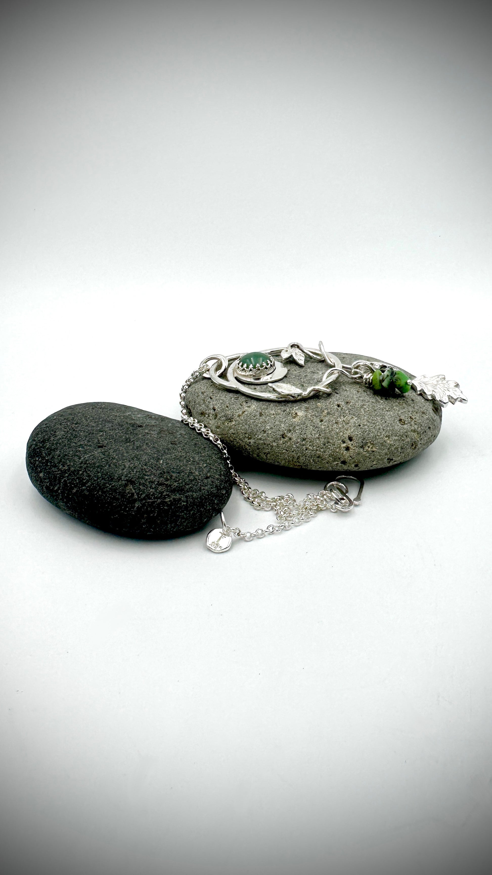 Sterling silver Dryad pendant laid flat on dark river stone with chain extended, showing botanical leaf and oak charm drops