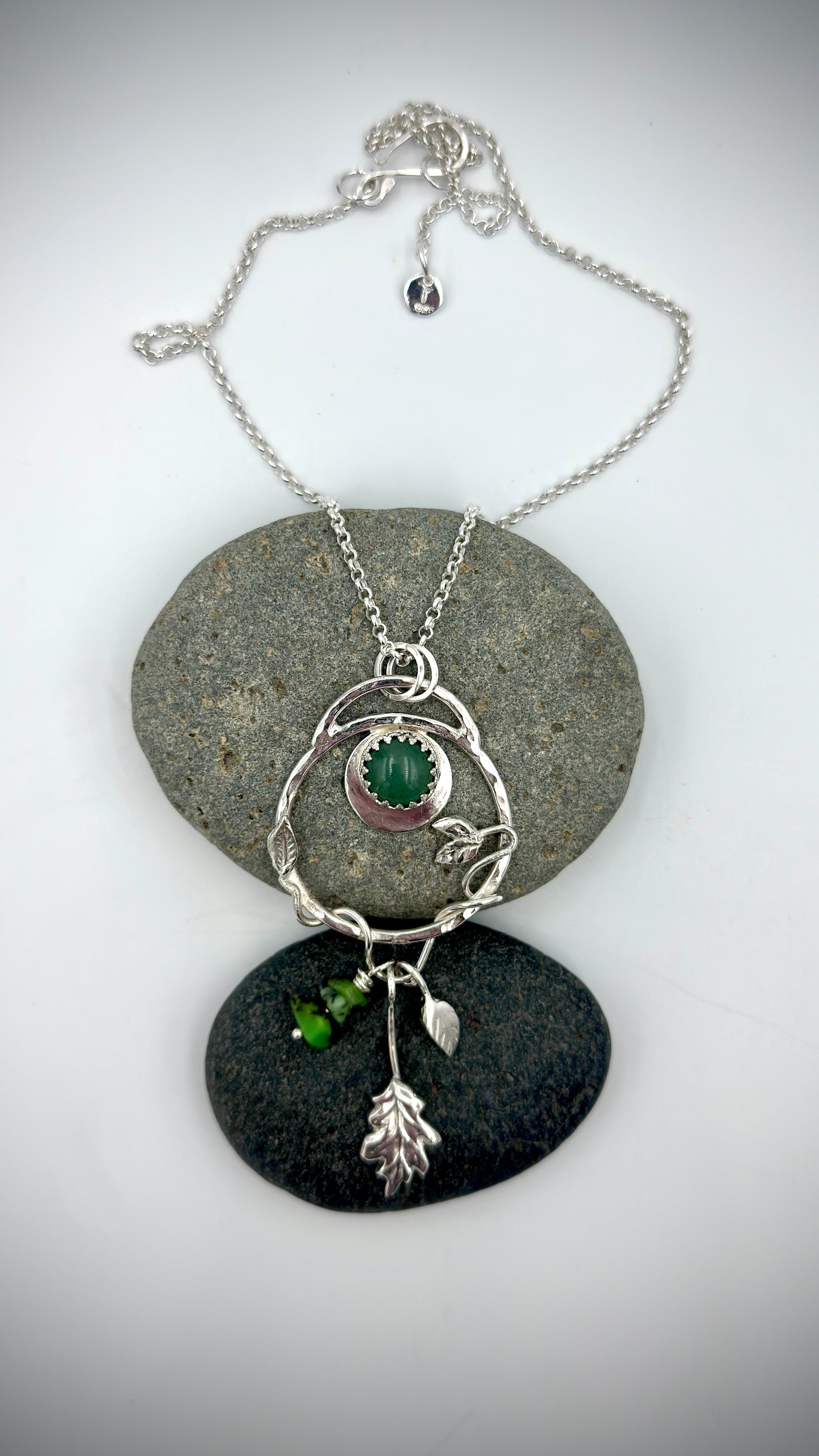 Sterling silver Dryad pendant with green aventurine cabochon, crescent moon setting, leaf and oak charm drops, displayed on natural river stones