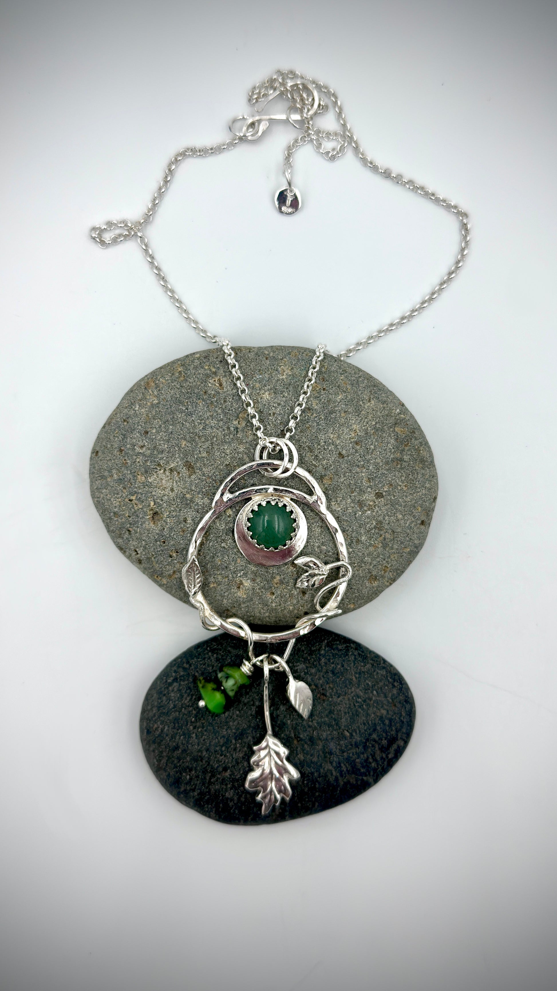 Sterling silver Dryad pendant with green aventurine cabochon, crescent moon setting, leaf and oak charm drops, displayed on natural river stones
