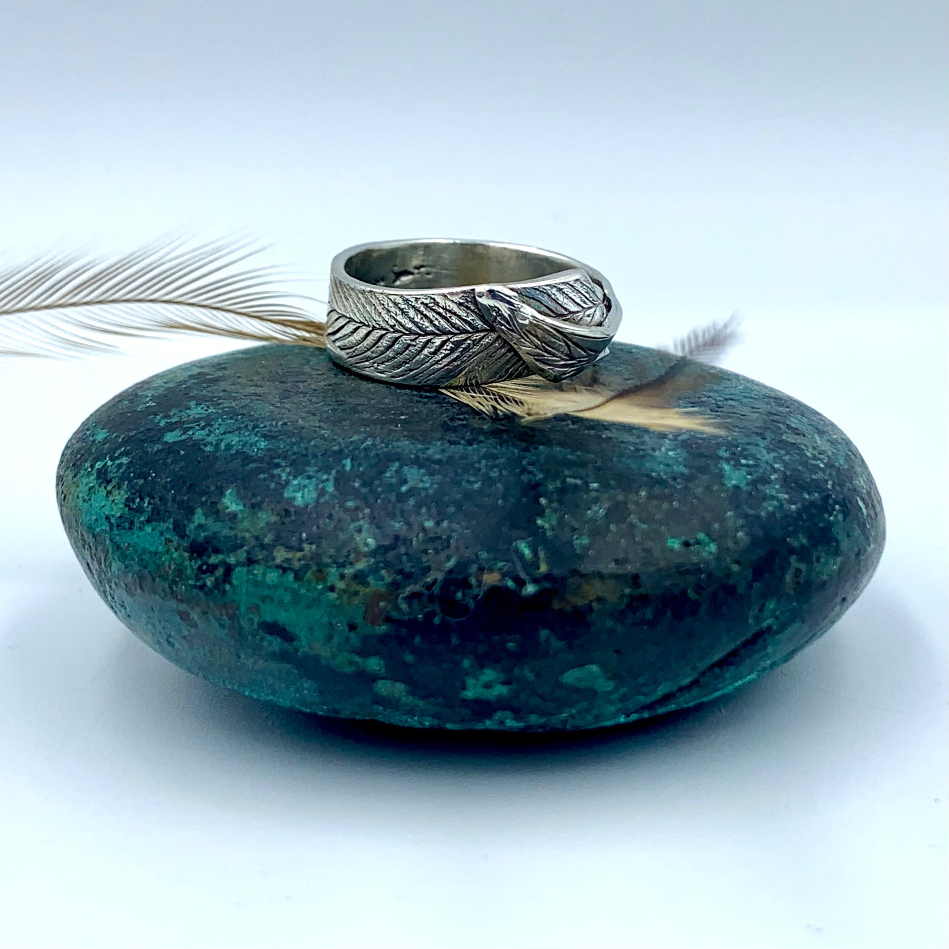Sterling silver emu feather imprinted ring, side view on a large dark green bronze patinated stone alongside a natural emu feather, showing full wrap feather texture and oxidised detail