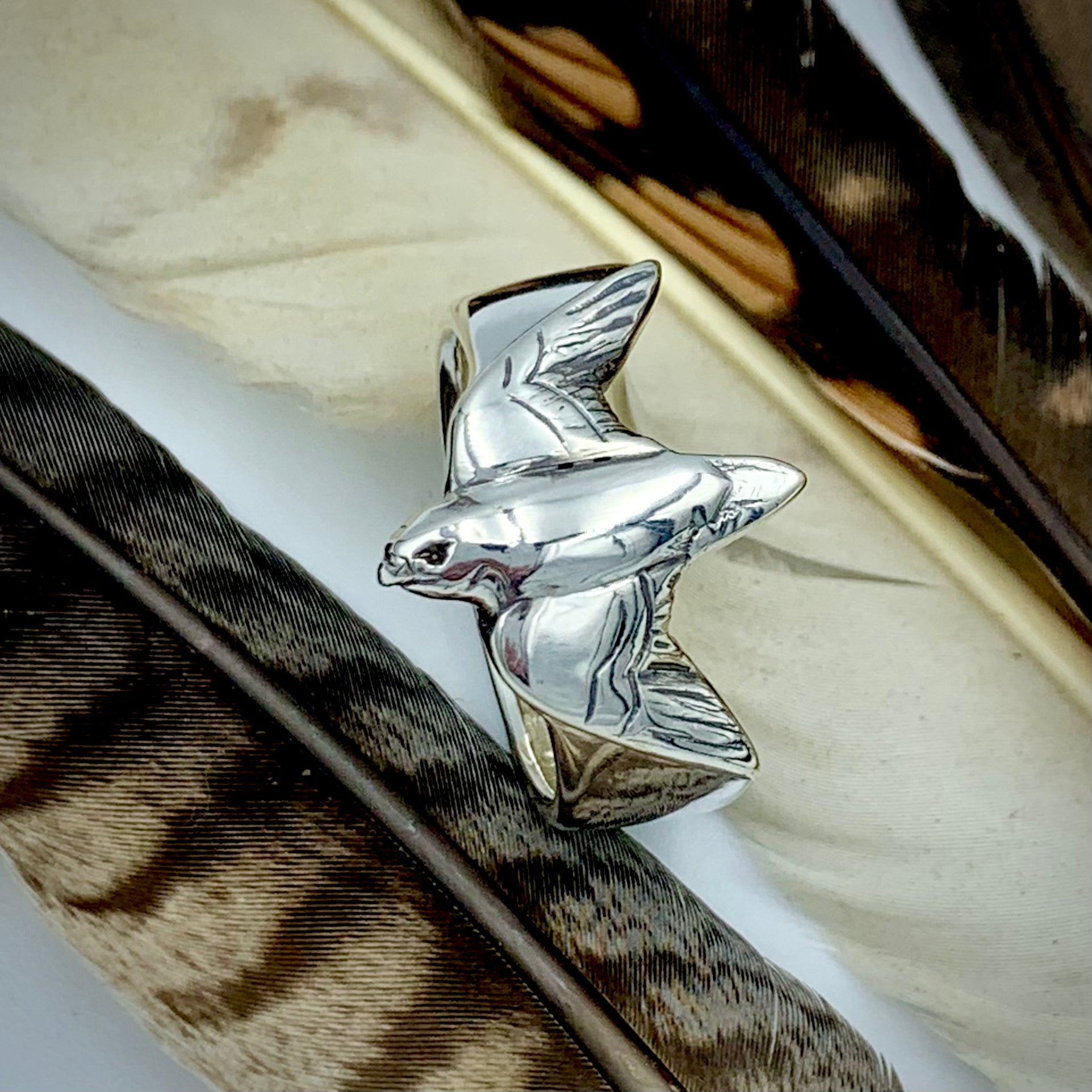 Sterling silver falconer's ring resting on a large brown and white raptor feather, top-down view showing sculpted bird in flight with detailed wing engraving