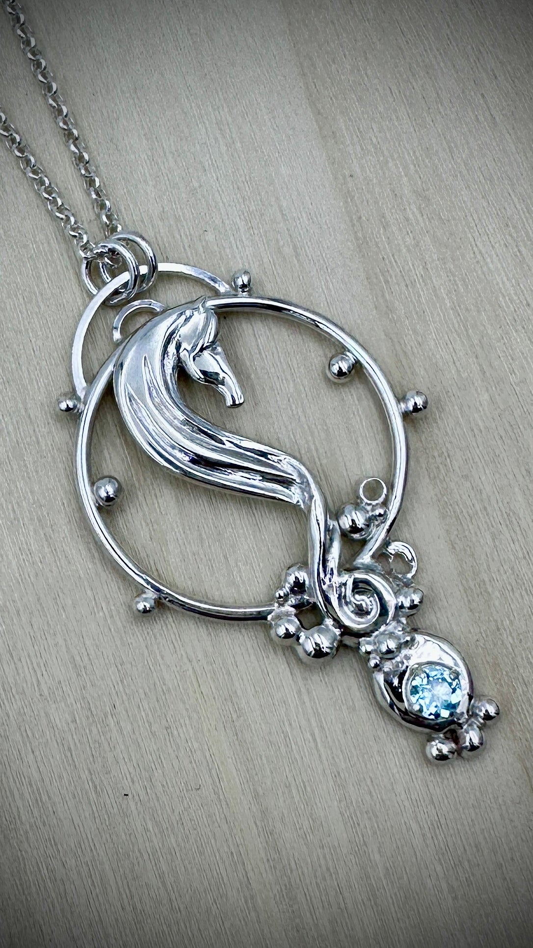 Sterling silver seahorse pendant, angled close-up on a light wood surface, highlighting the sculpted seahorse body, open circle frame with silver ball accents, scrollwork detail, and round blue topaz gemstone in a decorative bezel setting