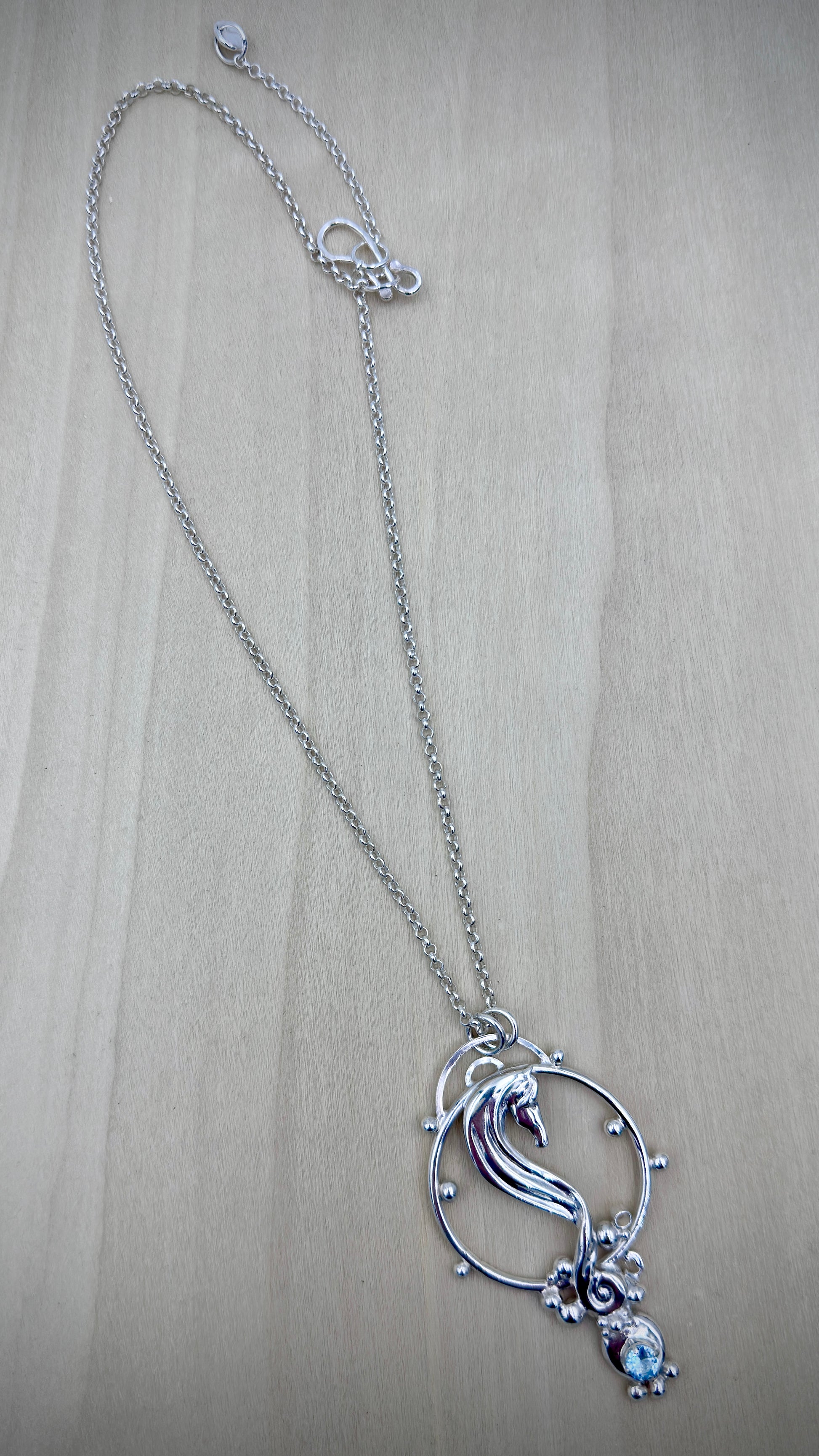 Sterling silver seahorse pendant necklace laid flat on a light wood surface, showing full chain length with hook clasp and extender, and the sculpted seahorse pendant with blue topaz drop