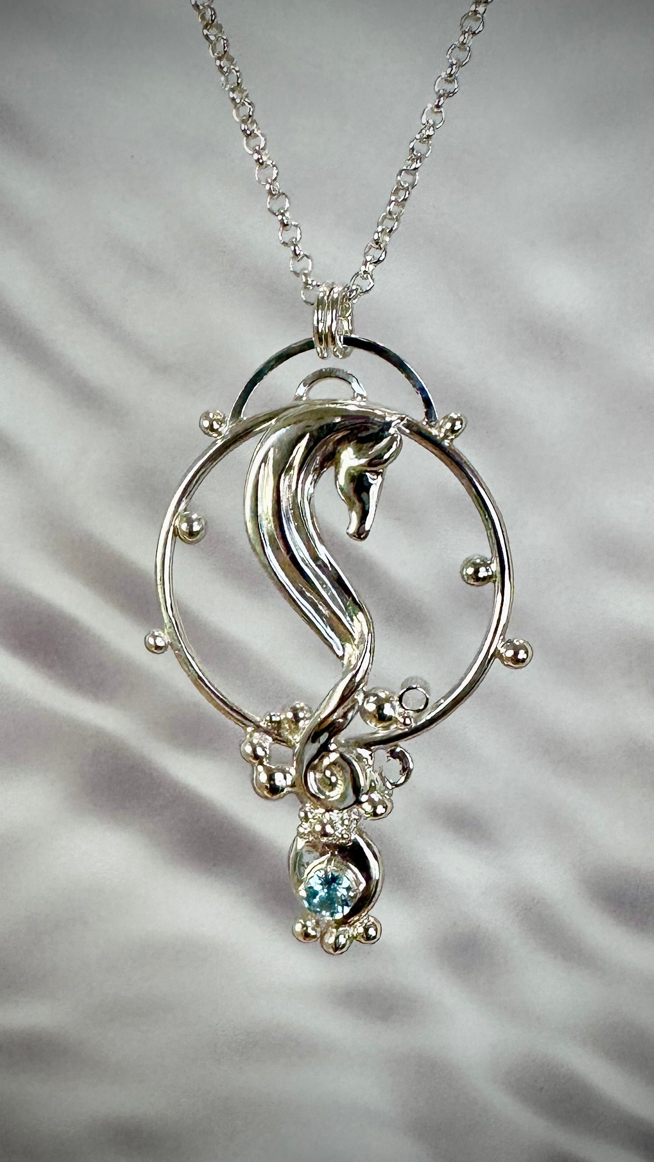 Sterling silver seahorse pendant necklace on a silver chain, front view against rippled background, featuring a sculpted seahorse within an open circle frame with silver ball accents and a round blue topaz gemstone drop