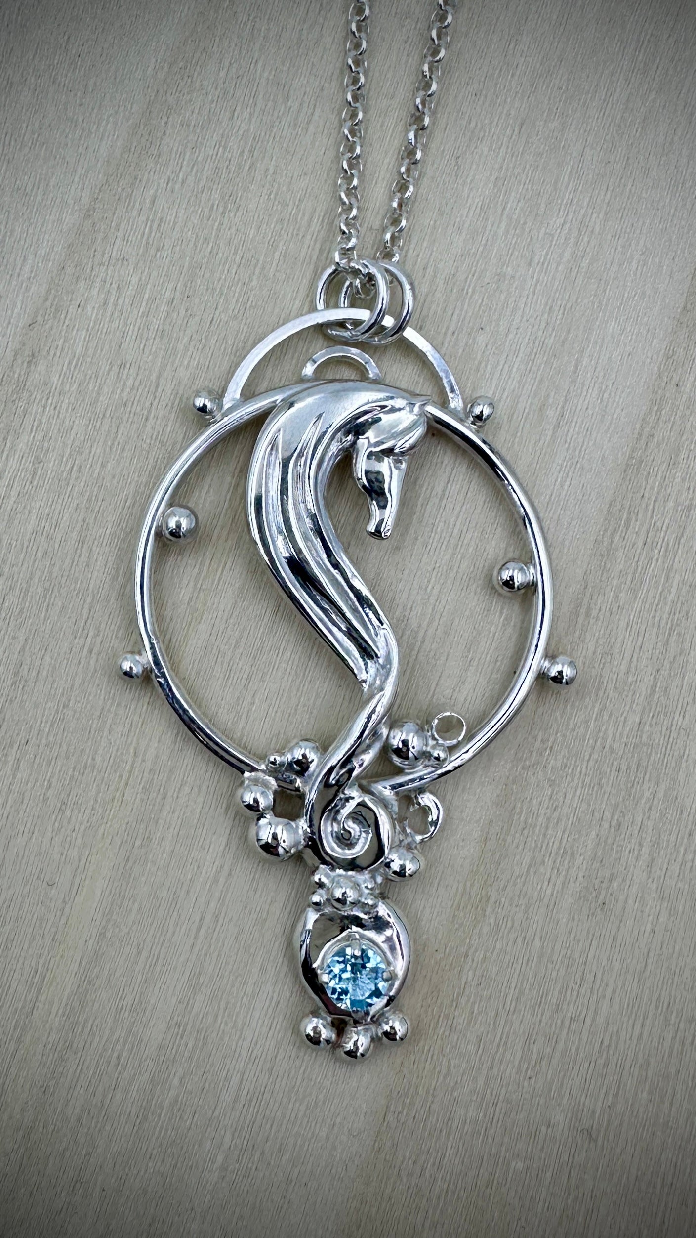 Sterling silver seahorse pendant necklace on a silver chain, front view on a light wood surface, showing sculpted horse within an open circular frame with silver ball details and a bezel-set round blue topaz drop