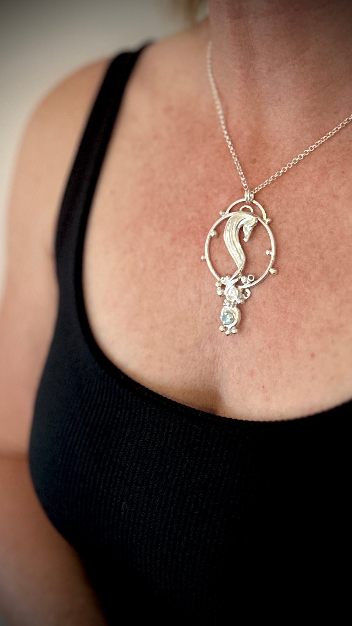Sterling silver seahorse pendant worn on a chain, shown against a black top, featuring a blue topaz gemstone drop and intricate scrollwork detail