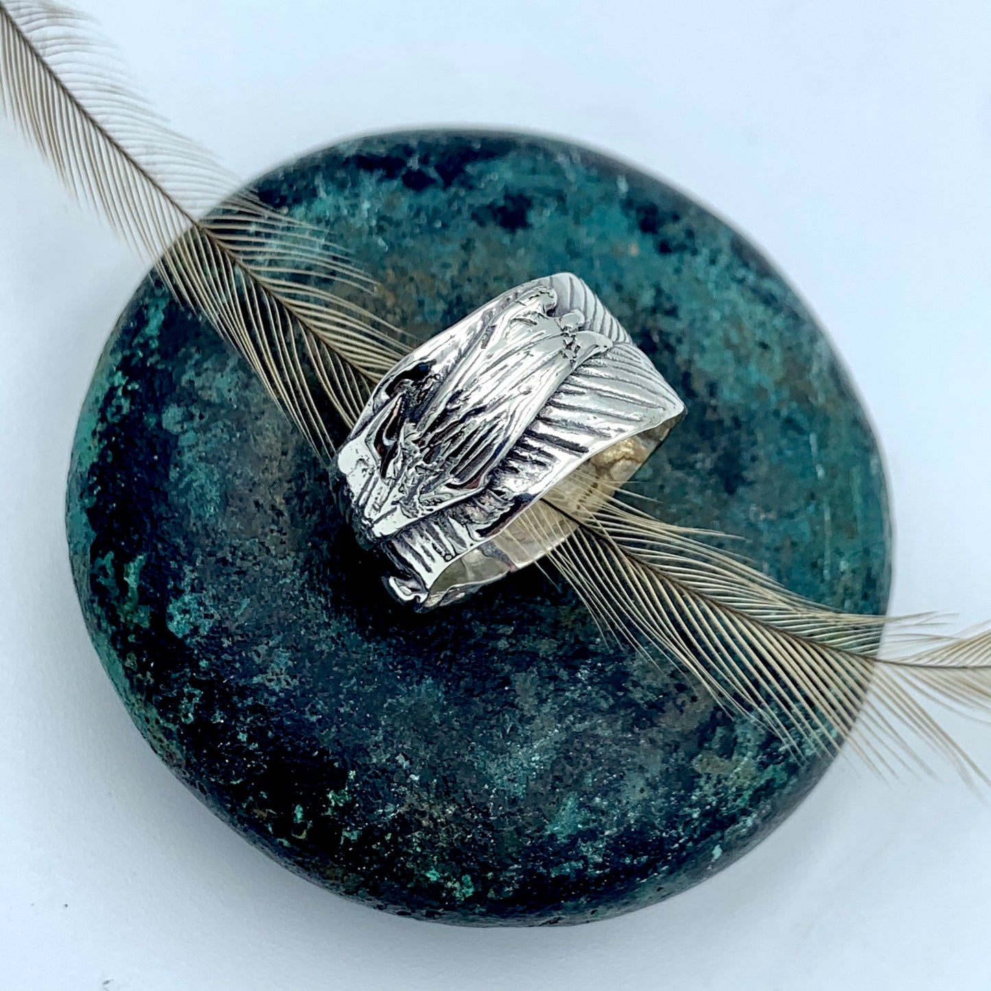 Sterling silver wide-band Australian emu feather ring, flat lay top view on a patinated green bronze stone with a natural emu feather, showing full feather imprint texture with oxidised shadow detail across the broad band