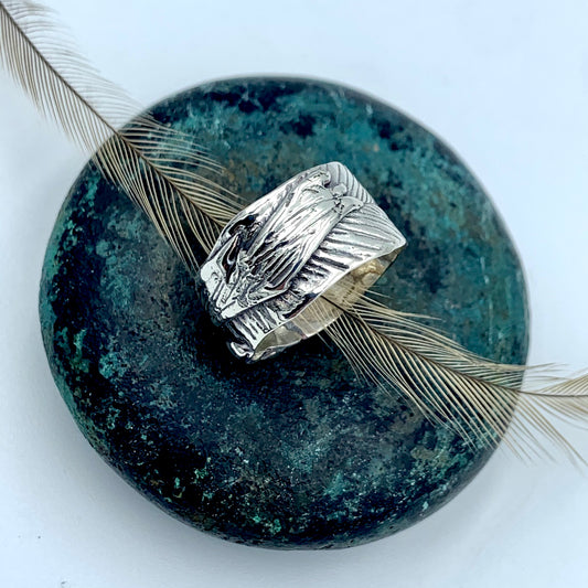 Sterling silver wide-band Australian emu feather ring, flat lay top view on a patinated green bronze stone with a natural emu feather, showing full feather imprint texture with oxidised shadow detail across the broad band