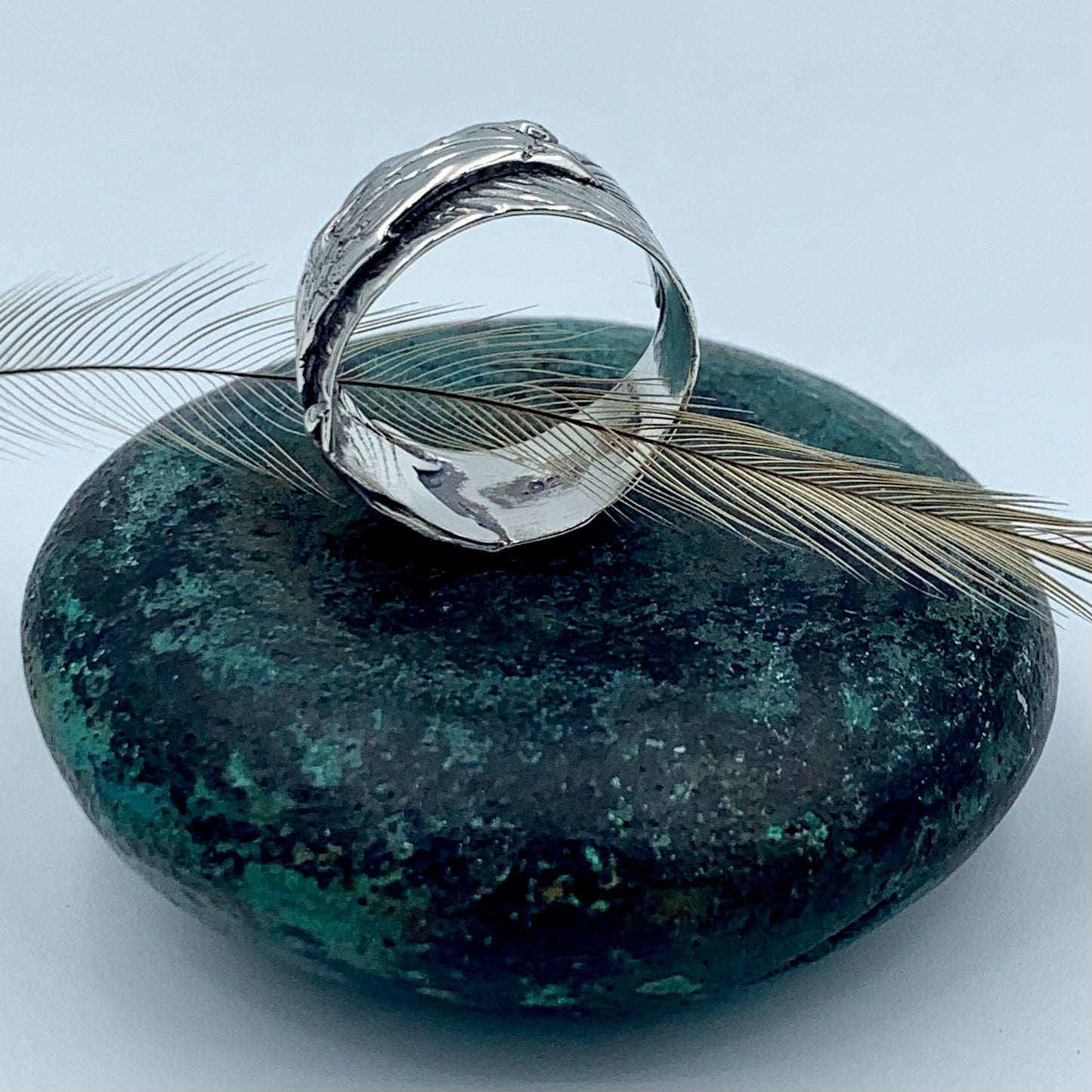 Sterling silver wide-band emu feather ring, side profile view on dark green stone with a natural emu feather, revealing polished interior and deeply textured oxidised feather exterior
