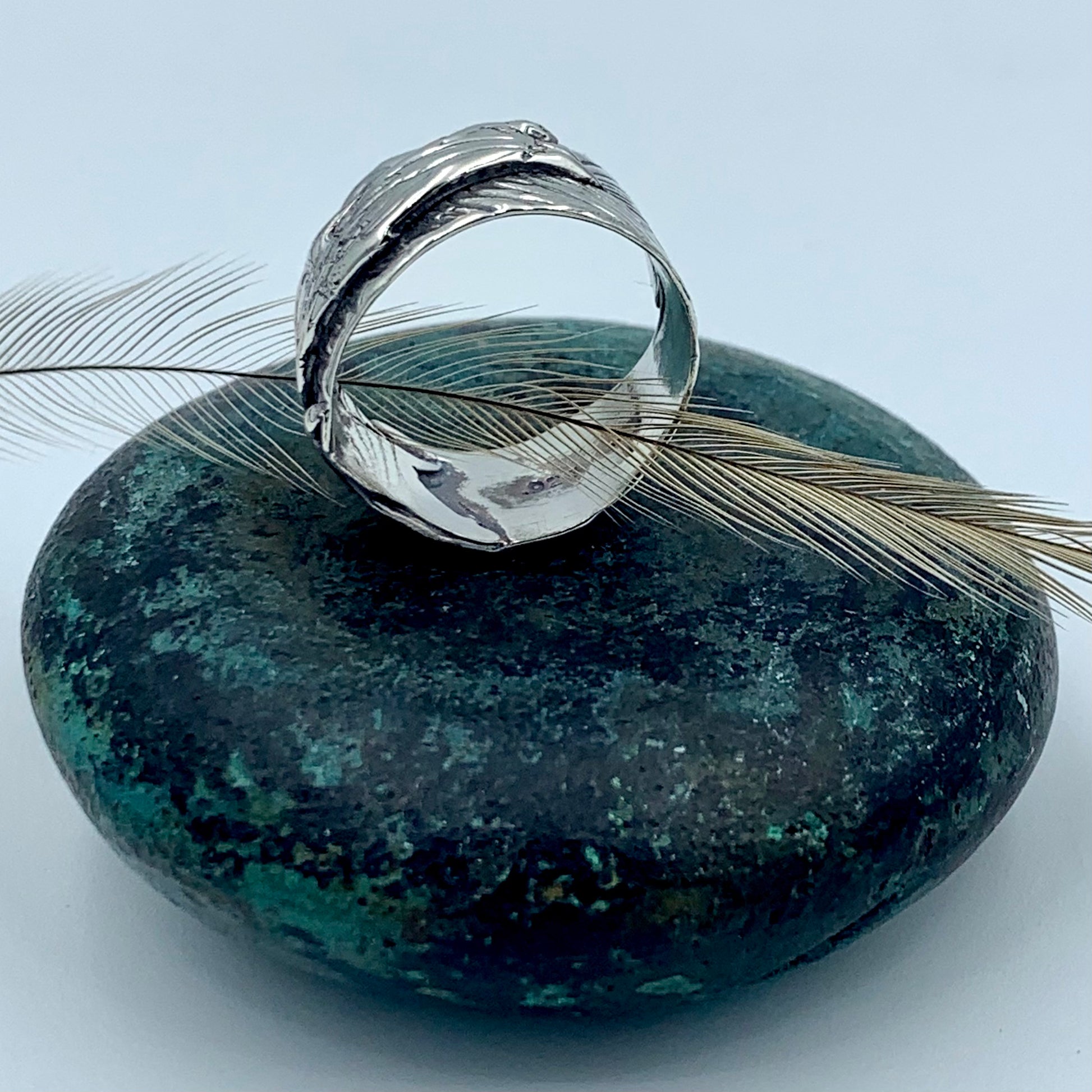 Sterling silver wide-band emu feather ring, side profile view on dark green stone with a natural emu feather, revealing polished interior and deeply textured oxidised feather exterior