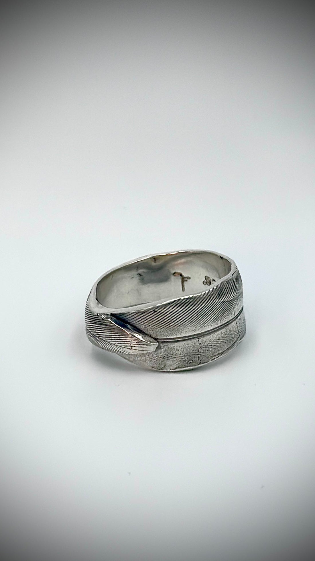 Three-quarter view of sterling silver bird's feather ring on white background, showing overlapping feather wrap and maker's marks inside the band — Marina Fox Fine Art
