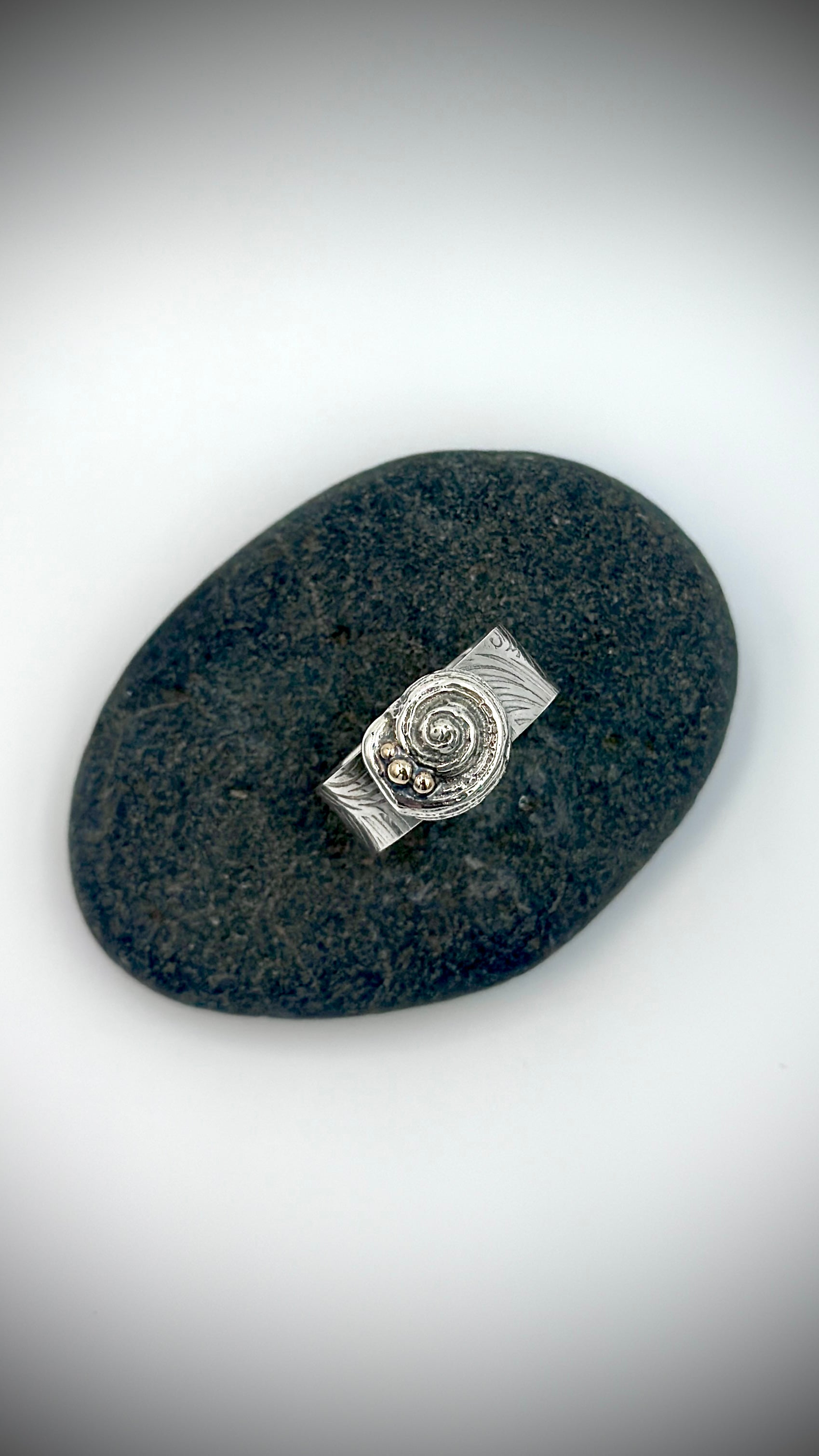Top-down view of sterling silver Beach Ring on dark river stone, spiral shell and granule detail centred on wave-patterned wide band — Marina Fox Fine Art