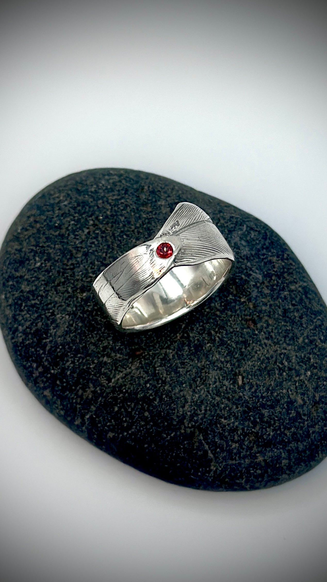 Top-down view of sterling silver bird's feather ring lying on dark river stone, red garnet centred at the feather quill, full feather texture visible — Marina Fox Fine Art