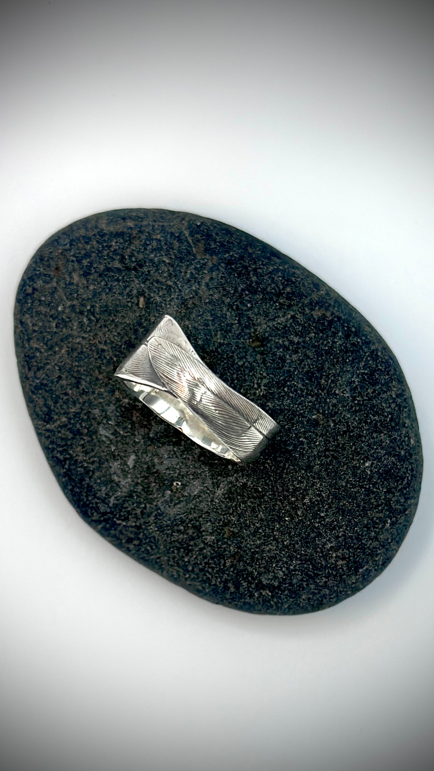 Top-down view of sterling silver bird's feather ring lying on dark river stone, showing full lost wax cast feather surface and overlapping quill tip — Marina Fox Fine Art
