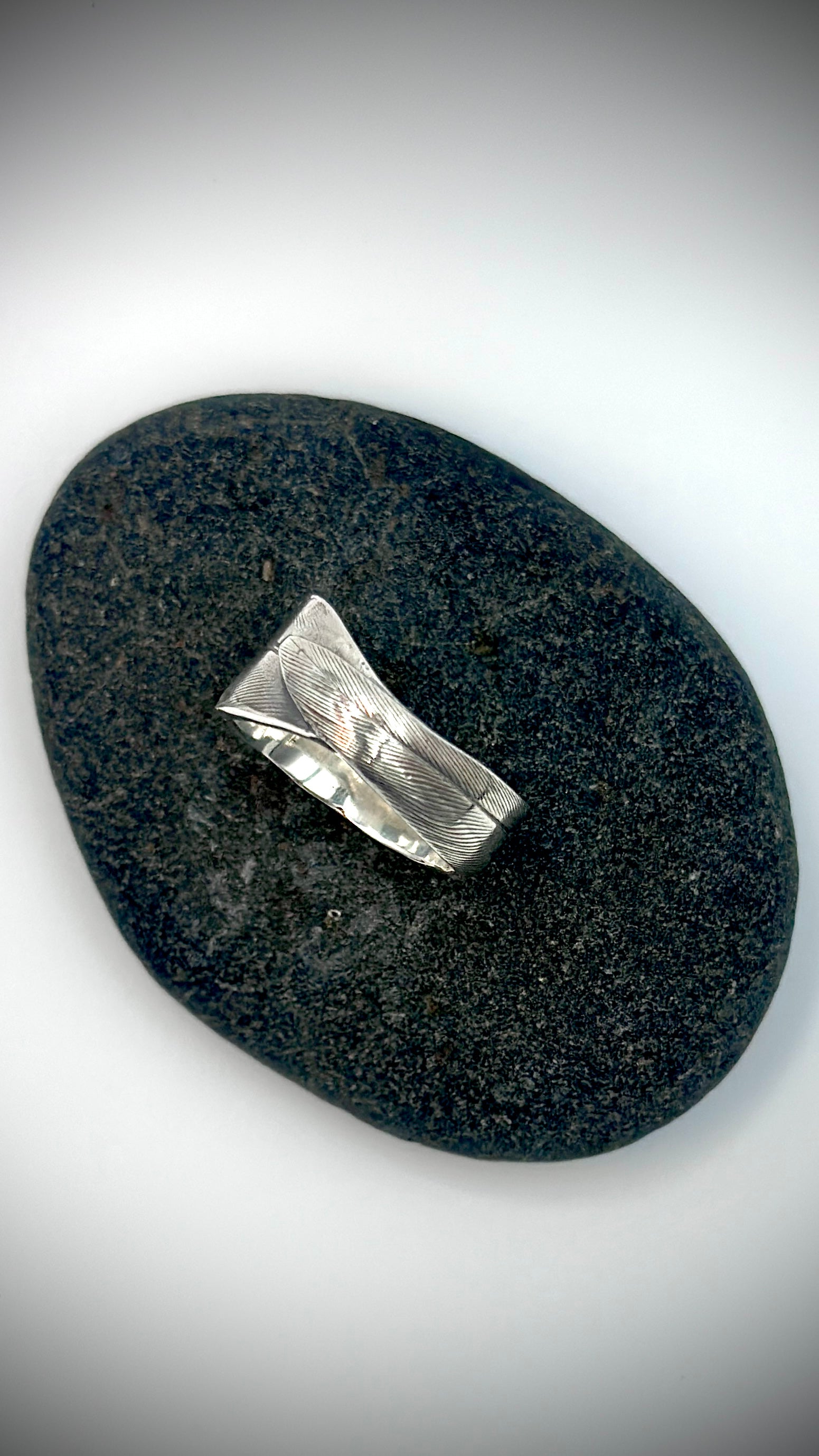 Top-down view of sterling silver bird's feather ring lying on dark river stone, showing full lost wax cast feather surface and overlapping quill tip — Marina Fox Fine Art