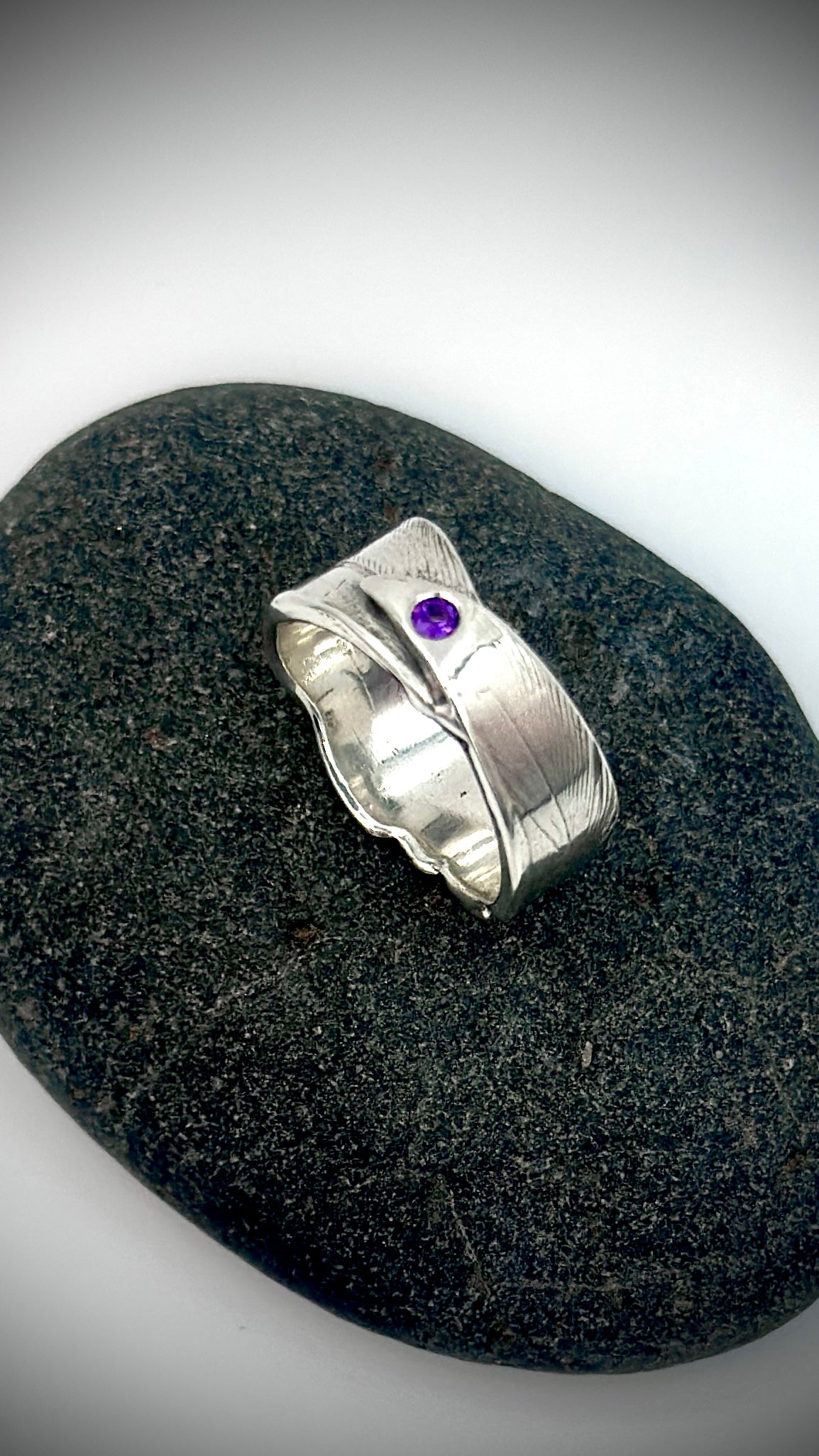 Top-down view of sterling silver bird's feather ring on dark river stone, purple amethyst centred on feather quill with organic wavy band edges — Marina Fox Fine Art