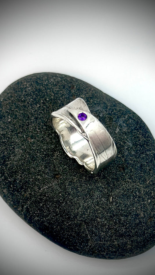 Top-down view of sterling silver bird's feather ring on dark river stone, purple amethyst centred on feather quill with organic wavy band edges — Marina Fox Fine Art