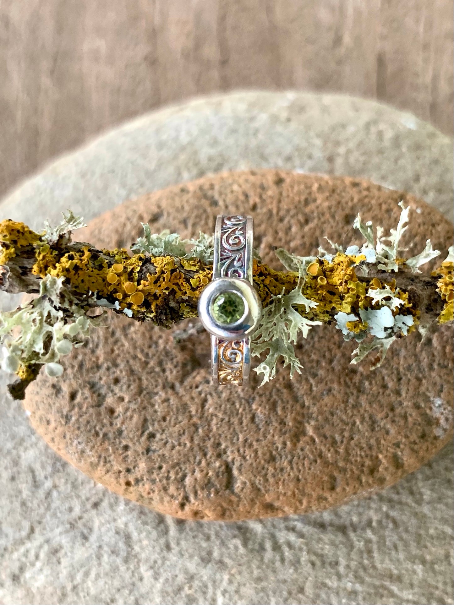 Top-down view of sterling silver peridot ring showing flush setting and scroll engraved band on natural stone