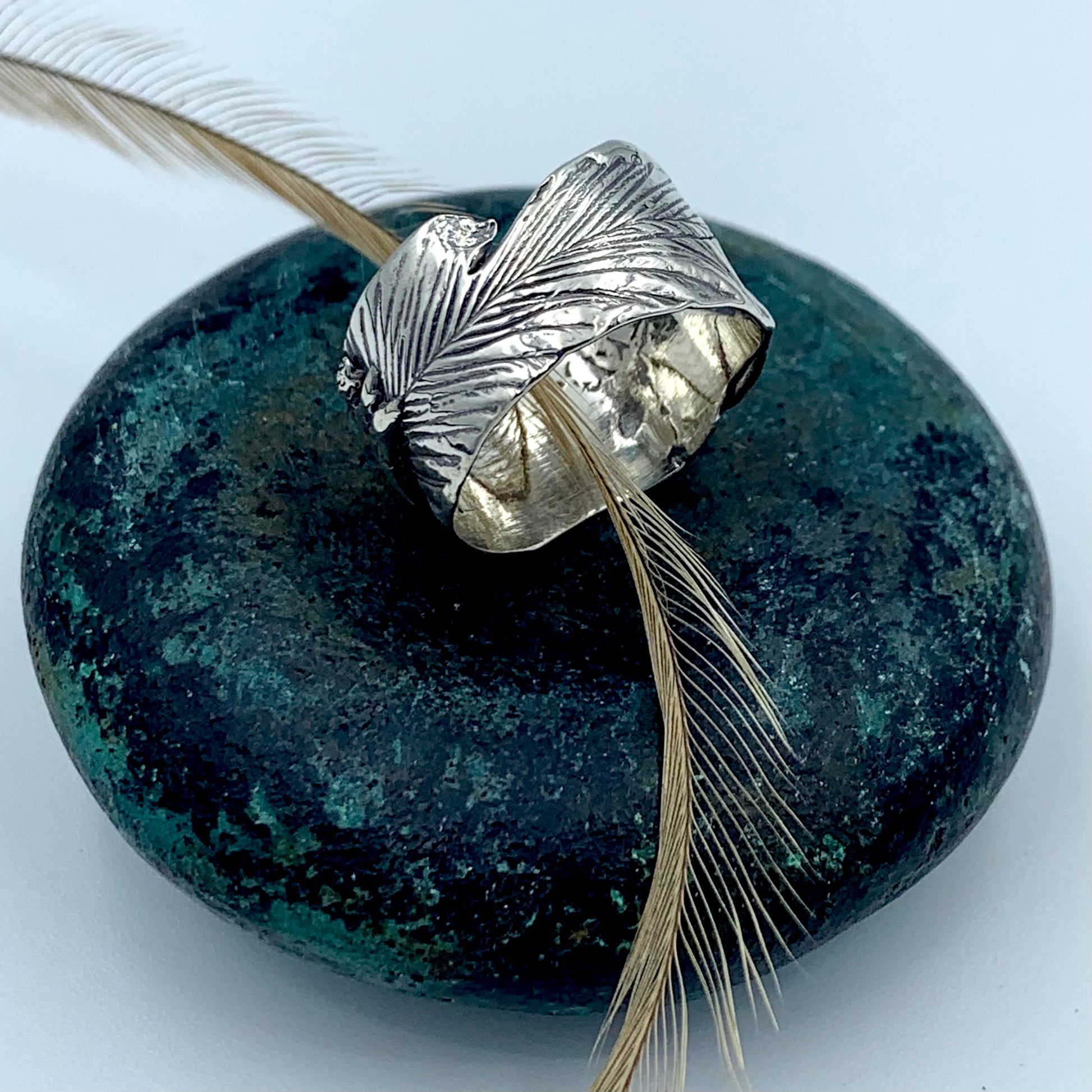 Wide sterling silver emu feather ring on patinated green bronze stone with a natural emu feather threaded through the band, showing sculptural feather texture and oxidised detailing across the broad surface - original Australian souvenir